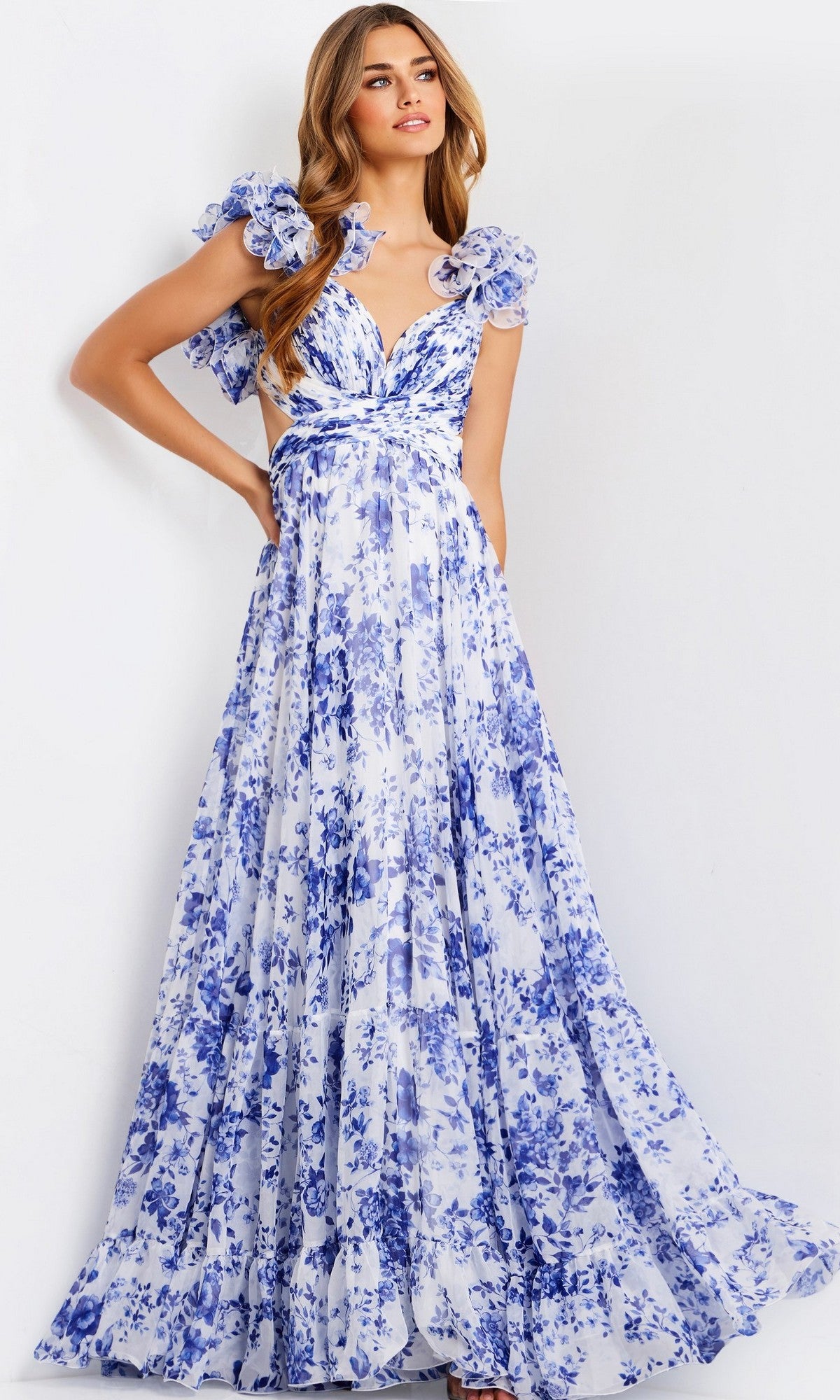 A woman models the Jovani 49143 Ruffled Long Print Prom Dress, featuring a floor-length blue and white floral design with ruffled sleeves and a deep V-neck, set against a plain white background.